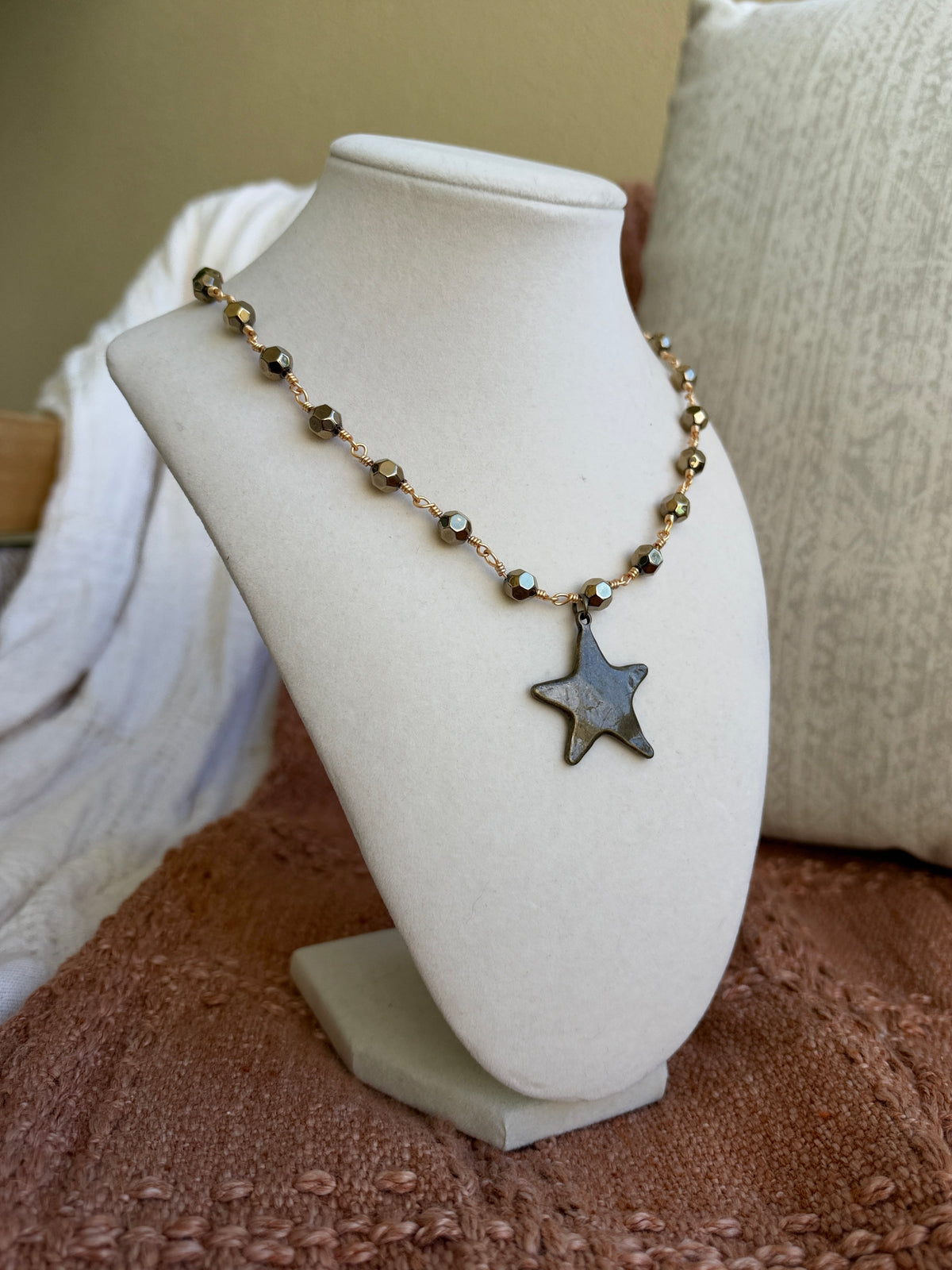 A beaded necklace with a star-shaped pendant displayed on a white jewelry bust. The necklace features metallic bronze beads and a dark gray star charm, set against a cozy indoor background with a textured brown blanket and neutral cushions.