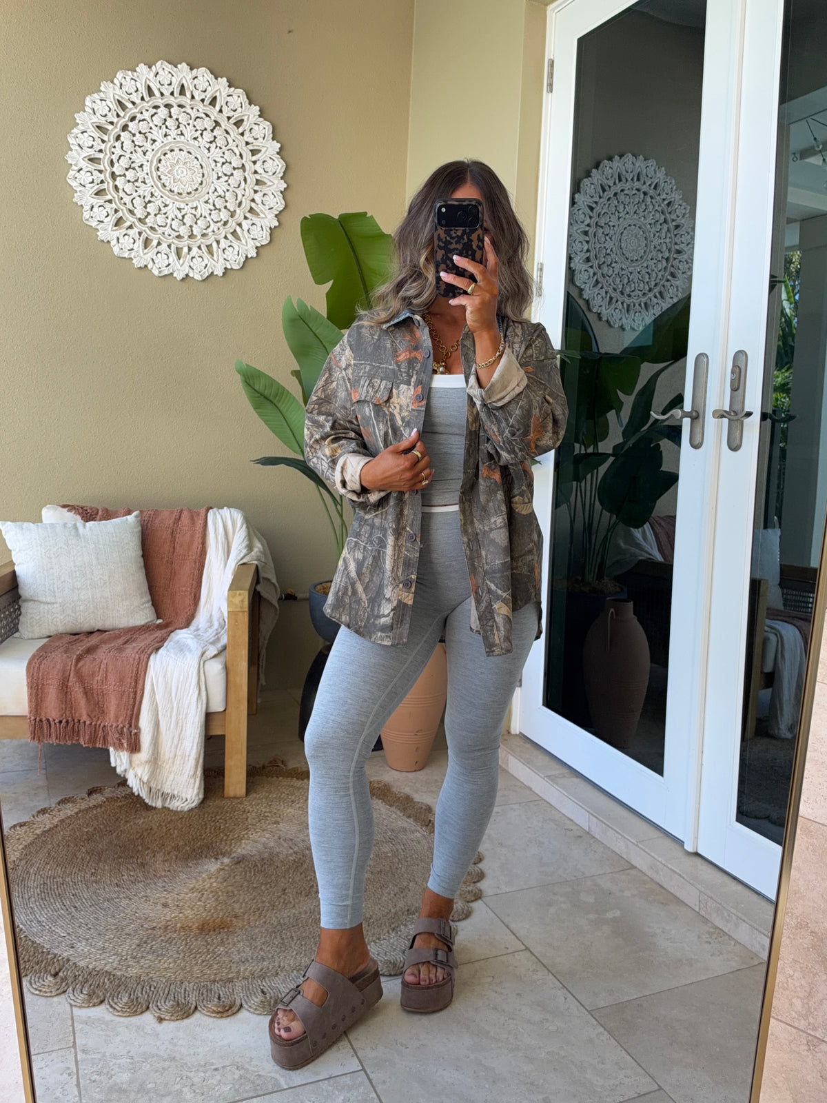 A woman takes a mirror selfie in a home setting wearing a brown and beige camouflage patterned button-up shirt over a white tank top, light gray leggings, and brown platform sandals.
