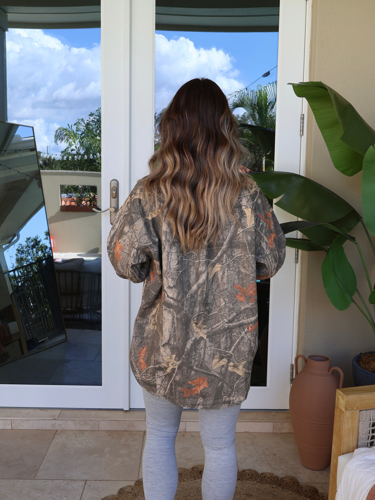 A woman viewed from behind wearing a camouflage hunting jacket with orange and brown details, standing in front of glass patio doors.