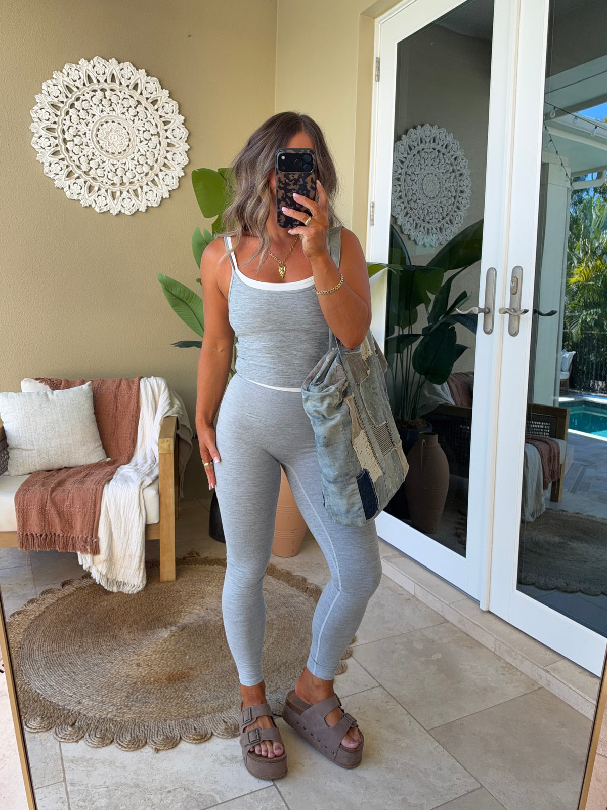 A woman takes a mirror selfie wearing a light gray ribbed tank top and matching high-waisted leggings with a white underband. She is holding a denim jacket and wearing brown platform sandals in a styled home setting with a beige wall, decorative wall hanging, and glass doors.