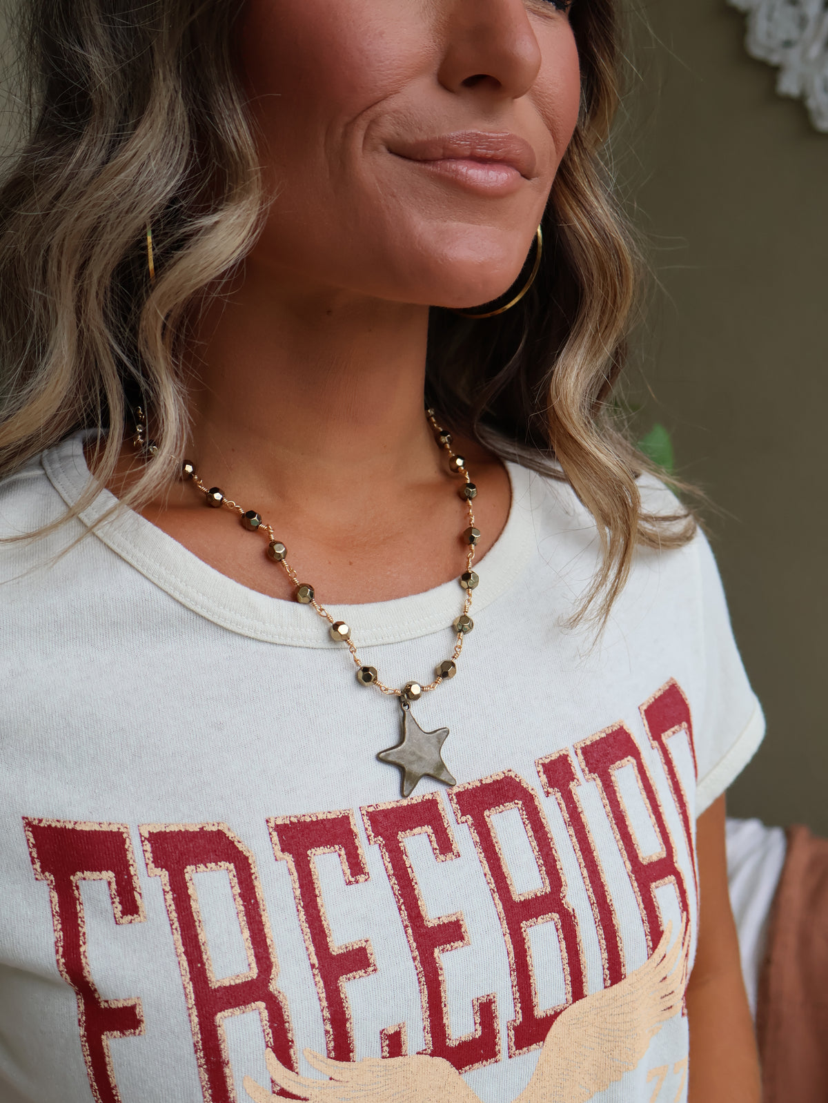 A close-up lifestyle shot of a woman wearing a white graphic t-shirt with distressed red lettering that reads 'FREEBIRD' and a silver star pendant necklace.