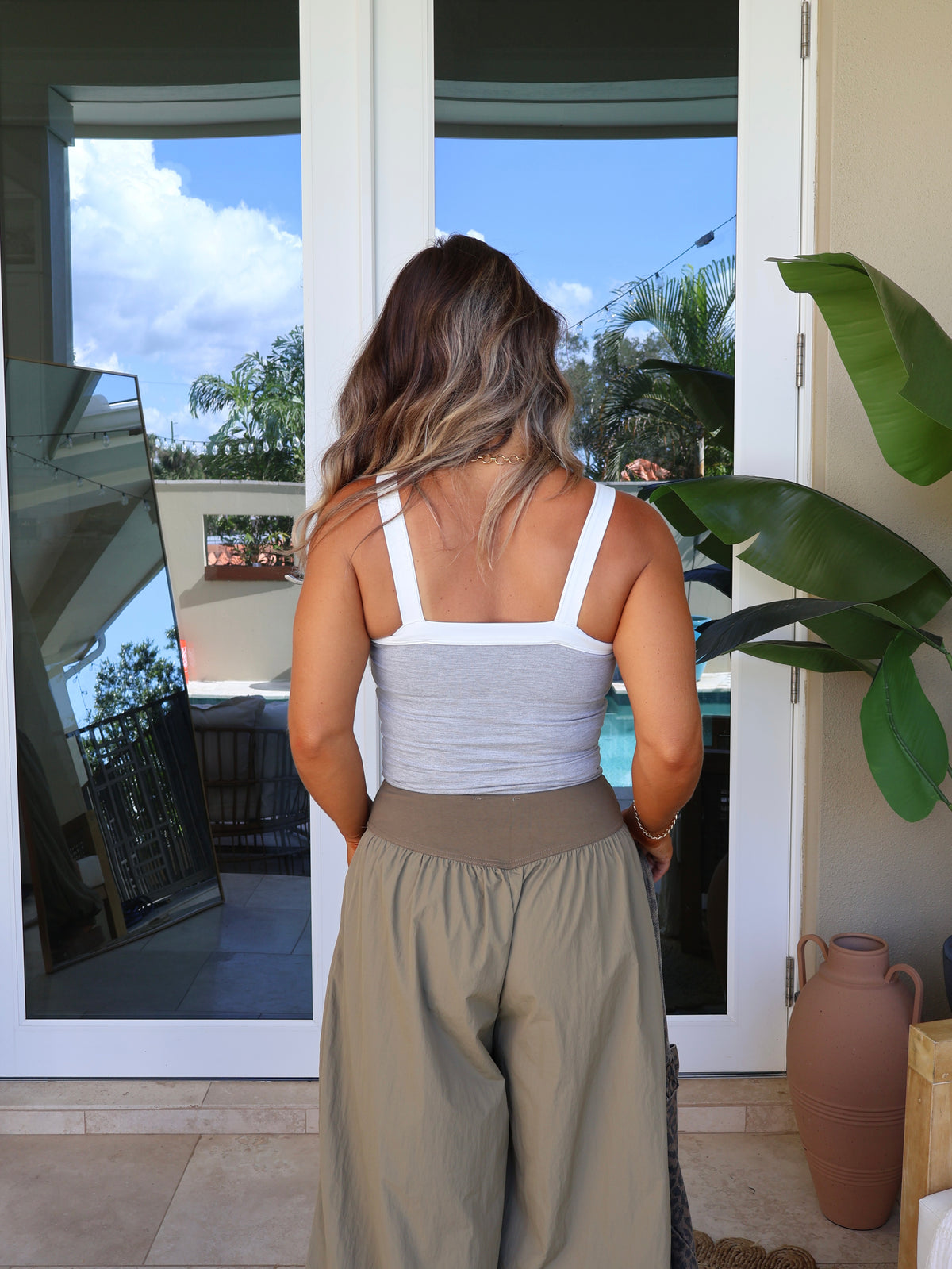 A woman viewed from behind wearing a white ribbed tank top and beige wide-leg pants, standing on a patio in front of glass doors with a pool and plants visible in the background.