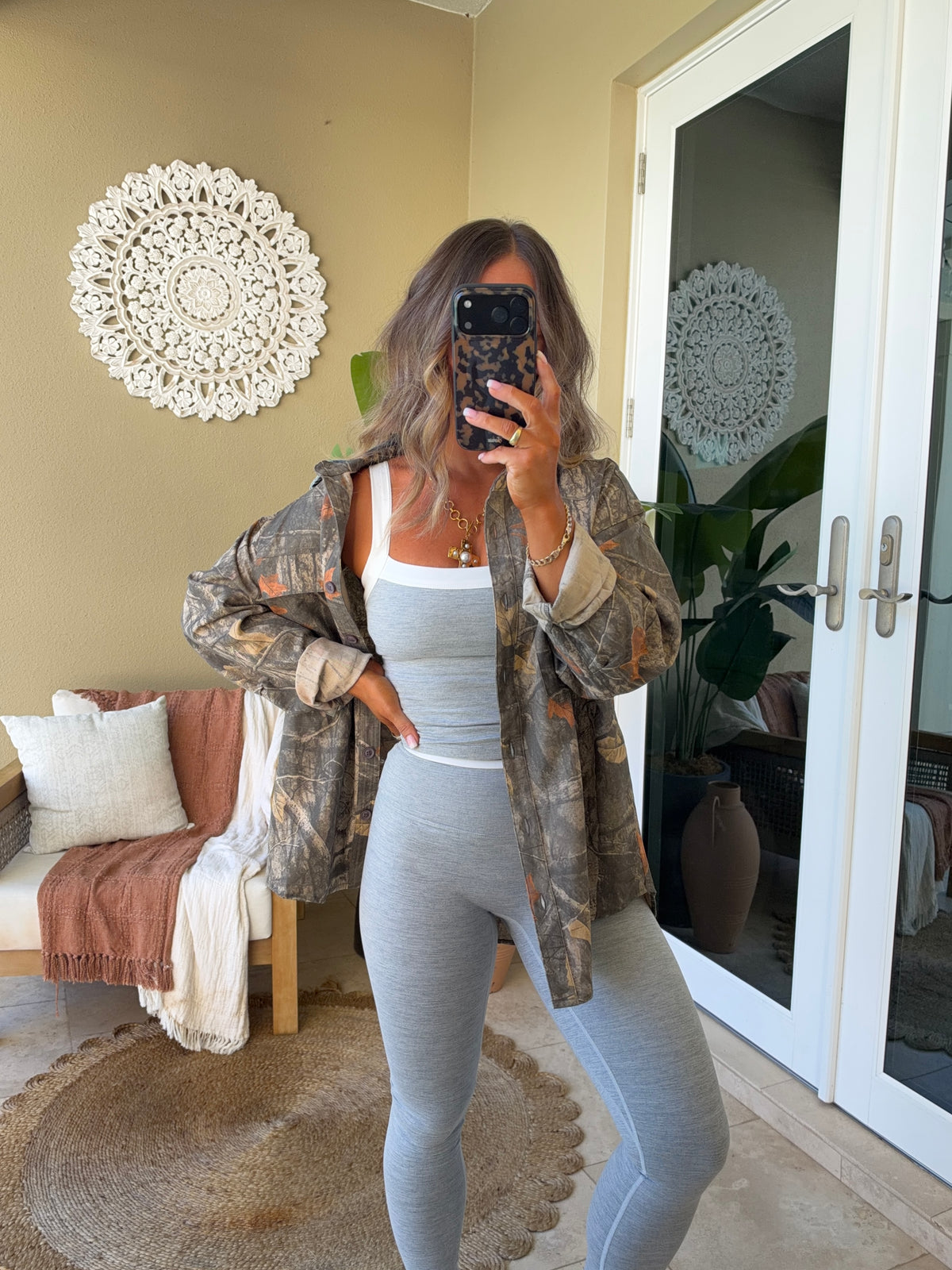 A woman takes a mirror selfie in a home setting, wearing a light gray ribbed tank top and matching high-waisted leggings, with a brown and beige camouflage jacket draped over her shoulders.