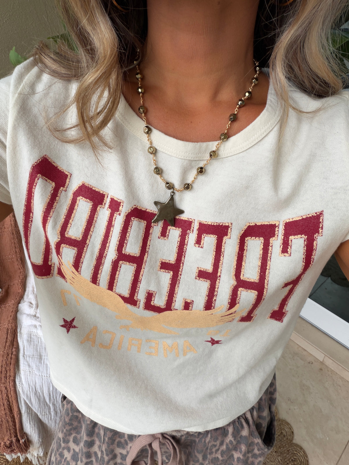A close-up of a person wearing a cream-colored graphic t-shirt with distressed red and gold lettering that reads 'FREE BIAD AMERICA'. The person is also wearing a beaded necklace with a star pendant.