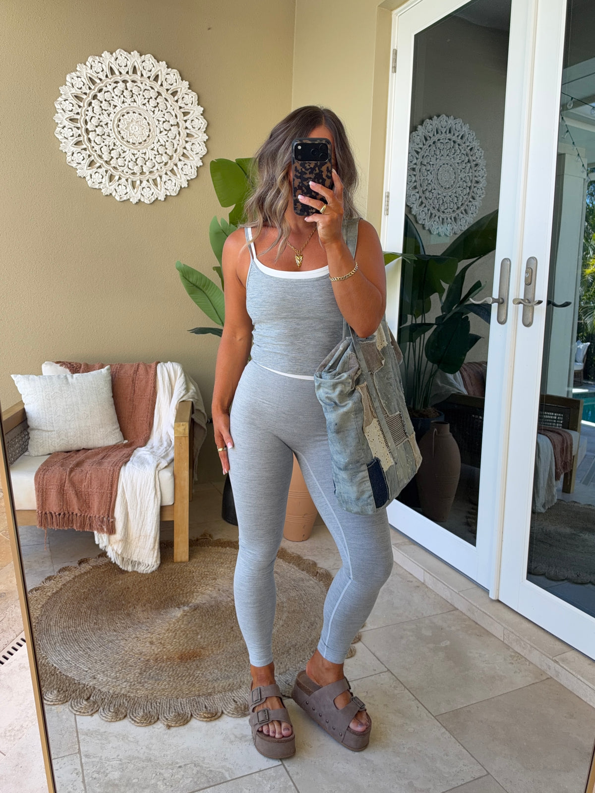 A woman takes a mirror selfie wearing a light gray ribbed tank top and matching high-waisted leggings, styled with a light blue denim jacket and taupe-brown platform sandals in a bright, modern room.
